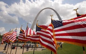 A row of large American Flags flap in the breeze with the Gateway Arch in the background