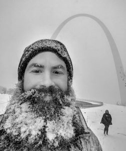 A man with a snowy beard poses in front of the Arch
