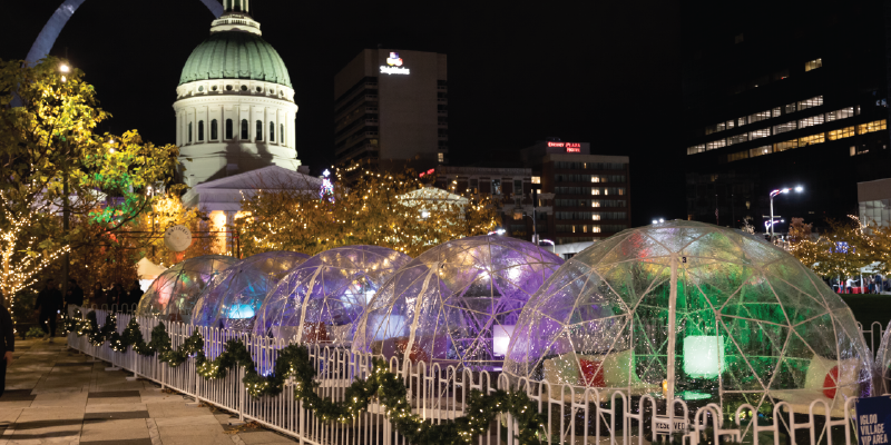 Winterfest private igloos are illuminated with colored lights and decorated for the holidays with a view of the Old Courthouse and Gateway Arch in the background.