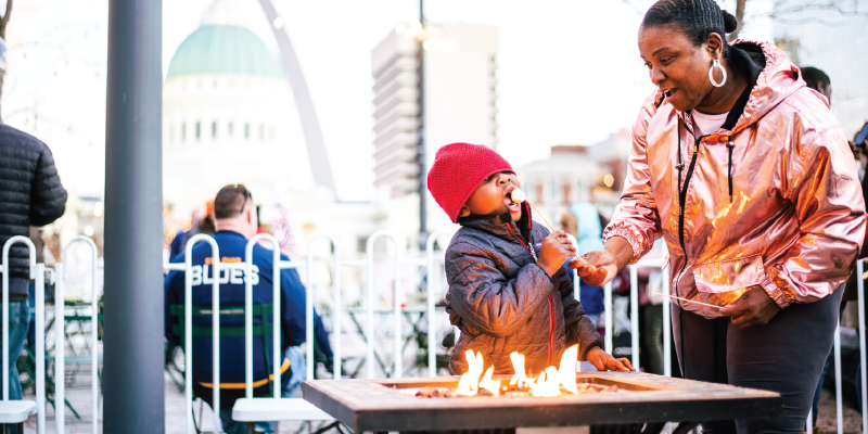 Young boy eating a roasted marshmallow off a stick with his mom at a S'mores station during Winterfest in Kiener Plaza.