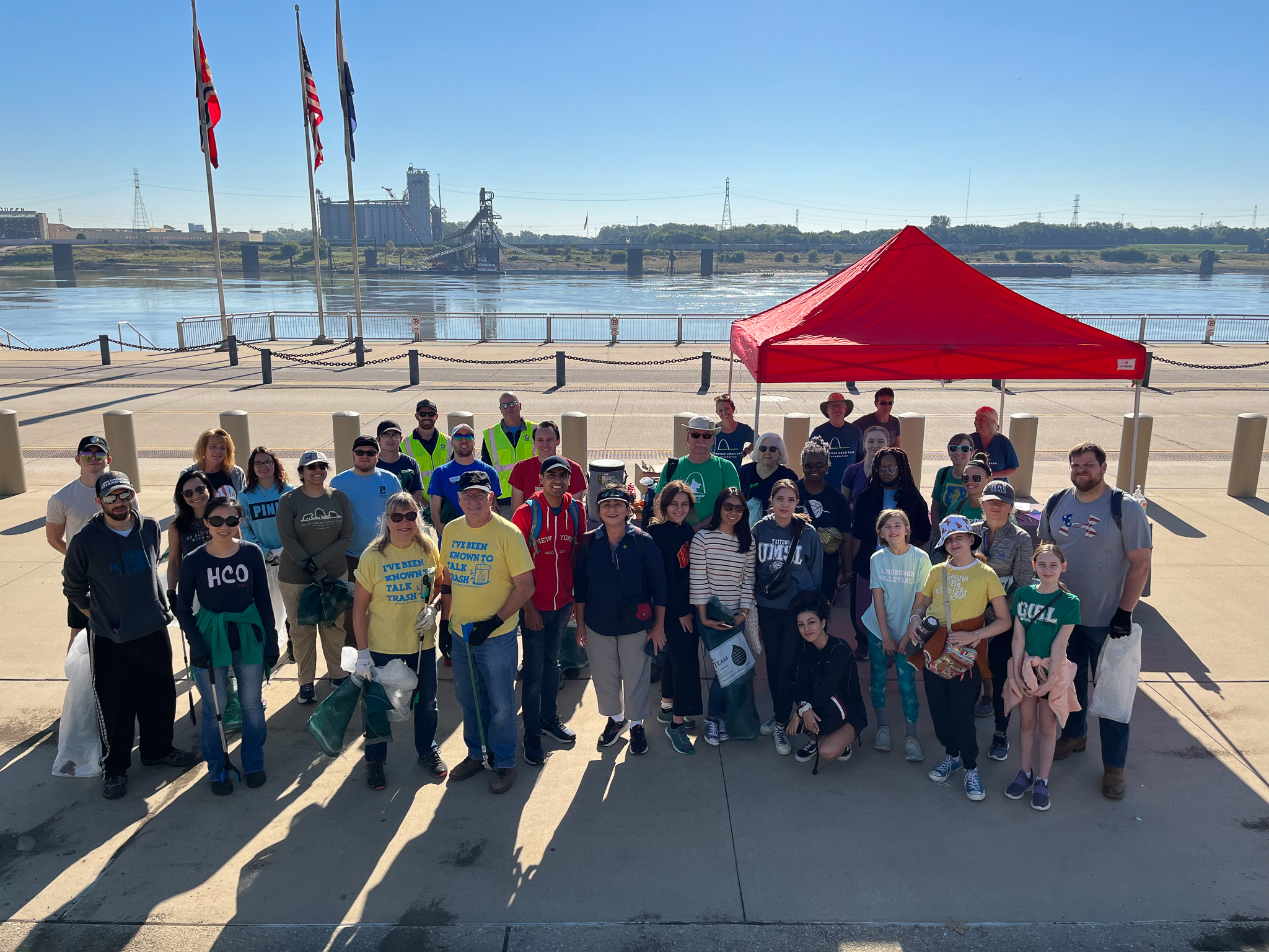 A group of about 2 dozen volunteers stand for a photo in front of the Mississippi River on the Gateway Arch National Park grounds with trash bags and pickers, ready to collect trash and clean up the St. Louis riverfront.